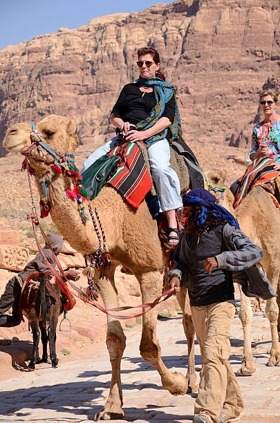 "Petra, Jordan - March 9th, 2012: Two mature Caucasian women riding camels between rocks of Petra. The first camel is led by a Bedouin. Boy with his donkey and rocks in the backgroud. Vertical image"