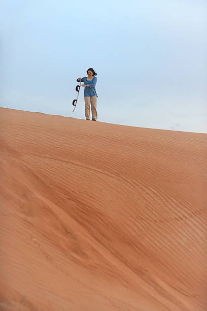 A happy, mature, Asian woman solo adventure tourist brushes sand off her sandboard while standing on top of a sand dune in the red desert between Dubai and Sharjah, United Arab Emirates.  Expansive, wide angle view with copyspace.