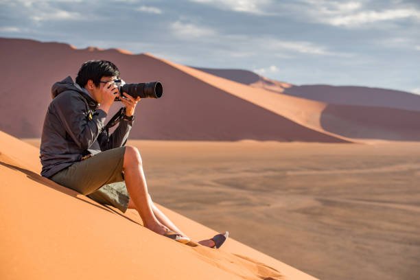 Young Asian man traveler and photographer sitting on sand dune taking photo of sunset in Namib desert of Namibia, Africa. Travel photography concept