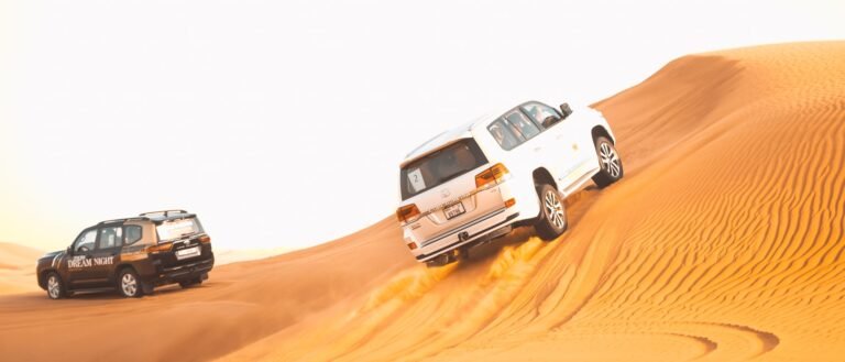 Two vehicles driving through the desert on a sand dune.