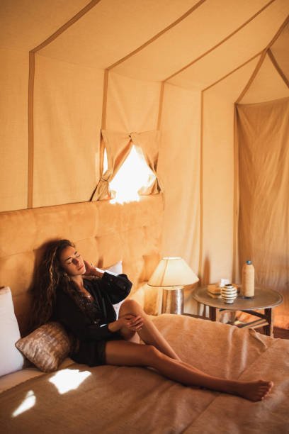 Sensual glamour portrait of Asian woman in interior of modern luxury glamping tent camp. Relaxing on bed. Eco design concept, natural and wooden elements in style.