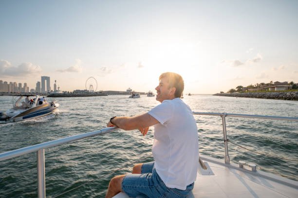 Adult man embraces the splendor of Dubai skyline as he siting on a yacht, captivated by the enchanting sunset over Dubai Marina. This tranquil moment exudes luxury and relaxation