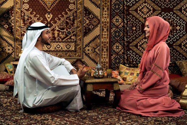 Young couple from Emirati spending time in an arabian traditional cafe. Man and woman wearing kandura and abaya from Dubai conversating together.