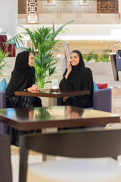 iStockalypse Dubai.  Two Emirati, Arabian young women wearing a traditional black abaya and hijab - one talking on her mobile phone - sitting in a modern lounge cafe of a luxury spa resort hotel.  Marble stairs, palm trees and elegant decorative railings on upper level in the background.  Dubai, UAE, Middle East, GCC.