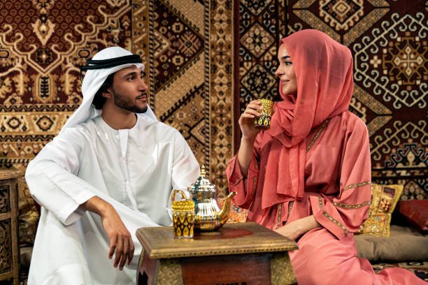 Young couple from Emirati spending time in an arabian traditional cafe. Man and woman wearing kandura and abaya from Dubai conversating together.